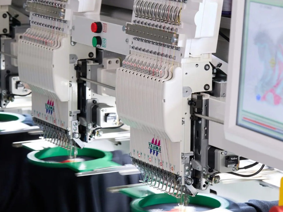 Row of Tajima embroidery heads stitching navy garments, the design previewed on the machine's screen