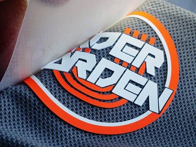 Raised silicone logo in orange and white, carrier film being peeled back from a grey textured jersey